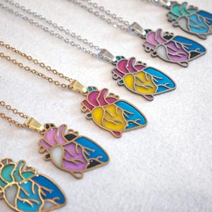 Könnte beinhalten: Eine Sammlung von herzförmigen Anhängerhalsketten. Jeder Anhänger zeigt ein farbenfrohes anatomisches Herzdesign mit rosa, gelben und blauen Akzenten. Die Halsketten haben entweder goldene oder silberne Ketten.