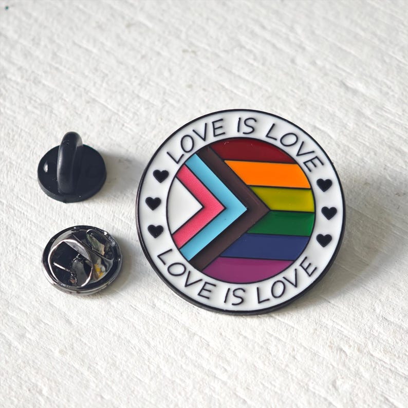 Puede incluir: Un pin redondo de esmalte con el dise&ntilde;o de la bandera Progress Pride, con franjas horizontales en naranja, amarillo, verde, azul y morado, y un chevr&oacute;n en blanco, rosa, azul claro, marr&oacute;n y negro. El borde blanco dice LOVE IS LOVE.