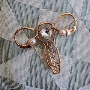 Puede incluir: Un broche de metal dorado con forma de &uacute;tero, que presenta un cristal transparente central en forma de coraz&oacute;n, dos perlas blancas redondas en los laterales y una fila vertical de cinco peque&ntilde;os cristales transparentes en la parte inferior.