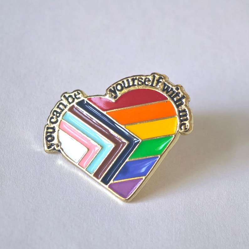 Puede incluir: Un pin de esmalte en forma de coraz&oacute;n con bordes de metal dorado. El dise&ntilde;o presenta los colores de la bandera Progress Pride, con franjas horizontales en rojo, naranja, amarillo, verde, azul y morado, y un chevr&oacute;n en blanco, rosa, azul claro, marr&oacute;n y negro. El texto "you can be yourself with me" est&aacute; grabado en negro en el borde superior.