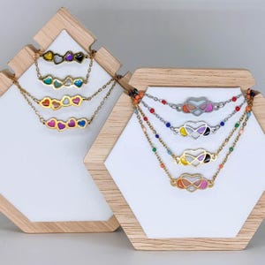 Könnte beinhalten: Fünf goldene Halsketten mit bunten Emaille-Herzen. Jede Halskette hat eine andere Farbkombination von Herzen. Die Halsketten werden auf einem Holzständer präsentiert.