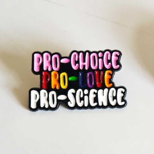 Puede incluir: Un pin de esmalte negro con tres l&iacute;neas de texto. La l&iacute;nea superior dice PRO-CHOICE en rosa, la l&iacute;nea central dice PRO-LOVE en rojo, amarillo, verde, azul y naranja, y la l&iacute;nea inferior dice PRO-SCIENCE en blanco. El pin tiene un borde ondulado.