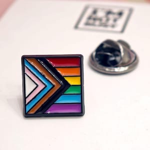 Puede incluir: Un pin cuadrado de esmalte con el dise&ntilde;o de la bandera Progress Pride. El pin muestra franjas horizontales de color rojo, naranja, amarillo, verde, azul y morado, con un patr&oacute;n de chevr&oacute;n a la izquierda en blanco, rosa, azul claro, marr&oacute;n y negro.