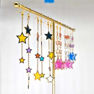 Peut inclure: Une présentation de boucles d'oreilles pendantes en forme d'étoile colorées avec des chaînes dorées. Les boucles d'oreilles sont disposées sur un support doré. Les étoiles sont remplies de différentes couleurs, notamment le jaune, le bleu, le rose, l'orange et le violet.