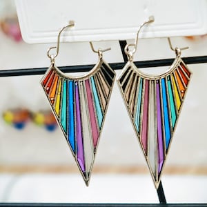 Può includere: Un paio di orecchini dorati con un design geometrico. Ogni orecchino presenta una forma triangolare con un arcobaleno di colori, tra cui rosso, arancione, giallo, verde, blu e viola. Gli orecchini hanno una chiusura a gancio.