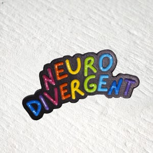 Puede incluir: Un pin de esmalte negro con la palabra NEURODIVERGENT en letras multicolores en relieve. Las letras est&aacute;n coloreadas en una secuencia de rosa, naranja, amarillo, verde lima, azul claro y morado, sobre un fondo negro oscuro y texturizado.