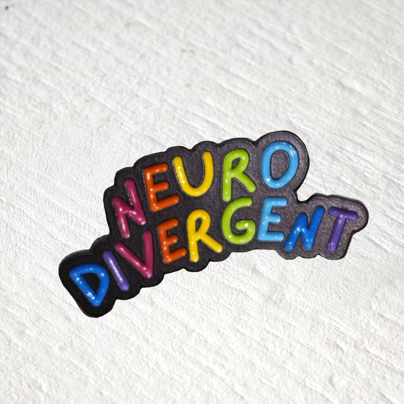 Puede incluir: Un pin de esmalte negro con la palabra NEURODIVERGENT en letras multicolores en relieve. Las letras est&aacute;n coloreadas en una secuencia de rosa, naranja, amarillo, verde lima, azul claro y morado, sobre un fondo negro oscuro y texturizado.