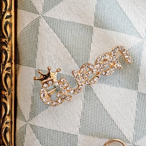 Puede incluir: Un broche de metal dorado con la forma de la palabra queen, que presenta una peque&ntilde;a corona sobre la letra q. Toda la superficie de las letras est&aacute; incrustada con peque&ntilde;os diamantes de imitaci&oacute;n transparentes y brillantes.