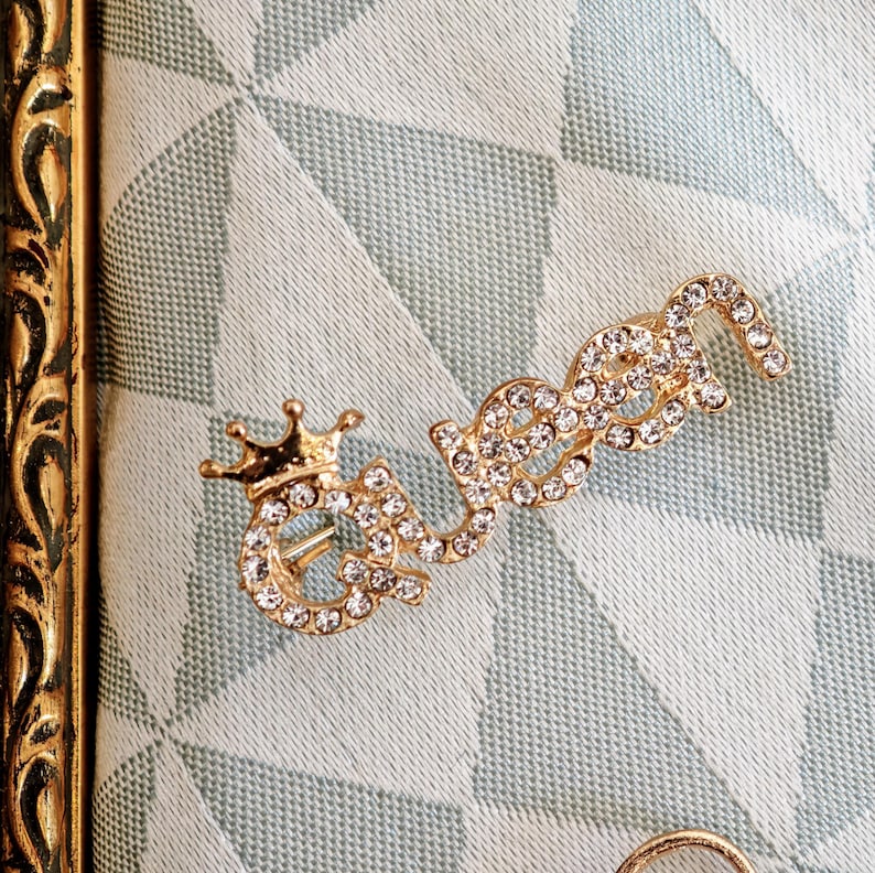 Puede incluir: Un broche de metal dorado con la forma de la palabra queen, que presenta una peque&ntilde;a corona sobre la letra q. Toda la superficie de las letras est&aacute; incrustada con peque&ntilde;os diamantes de imitaci&oacute;n transparentes y brillantes.