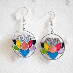 Könnte beinhalten: Ein Paar Ohrringe mit einem farbenfrohen Lotusblumen-Design. Jeder Ohrring hat einen silberfarbenen kreisförmigen Rahmen mit einem Lotus im Glasmalerei-Stil. Die Lotusblütenblätter sind in Rot-, Gelb-, Blau-, Rosa- und Lilatönen gehalten. Die Ohrringe hängen an silbernen Haken.