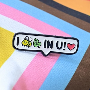Puede incluir: Un pin de esmalte blanco rectangular con borde negro que presenta el texto I, un icono de abeja amarilla y negra, un icono de hoja verde, el texto IN U! y un peque&ntilde;o icono de coraz&oacute;n rojo. El pin tiene forma de burbuja de di&aacute;logo.