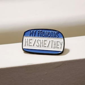 Puede incluir: Un pin de esmalte rectangular con forma de etiqueta de nombre con un borde azul y un centro blanco. El texto MY PRONOUNS est&aacute; impreso en negro en la secci&oacute;n superior azul, y HE/SHE/THEY est&aacute; impreso en negro en la secci&oacute;n central blanca.