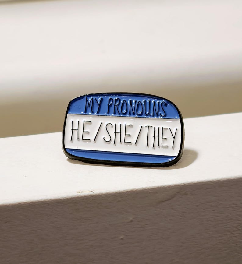 Puede incluir: Un pin de esmalte rectangular con forma de etiqueta de nombre con un borde azul y un centro blanco. El texto MY PRONOUNS est&aacute; impreso en negro en la secci&oacute;n superior azul, y HE/SHE/THEY est&aacute; impreso en negro en la secci&oacute;n central blanca.