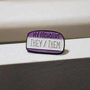Puede incluir: Un pin de esmalte de forma ovalada con un borde negro. La secci&oacute;n superior es morada con el texto MY PRONOUNS en negro. La secci&oacute;n inferior es blanca con el texto THEY / THEM en negro, imitando la apariencia de una etiqueta de nombre.