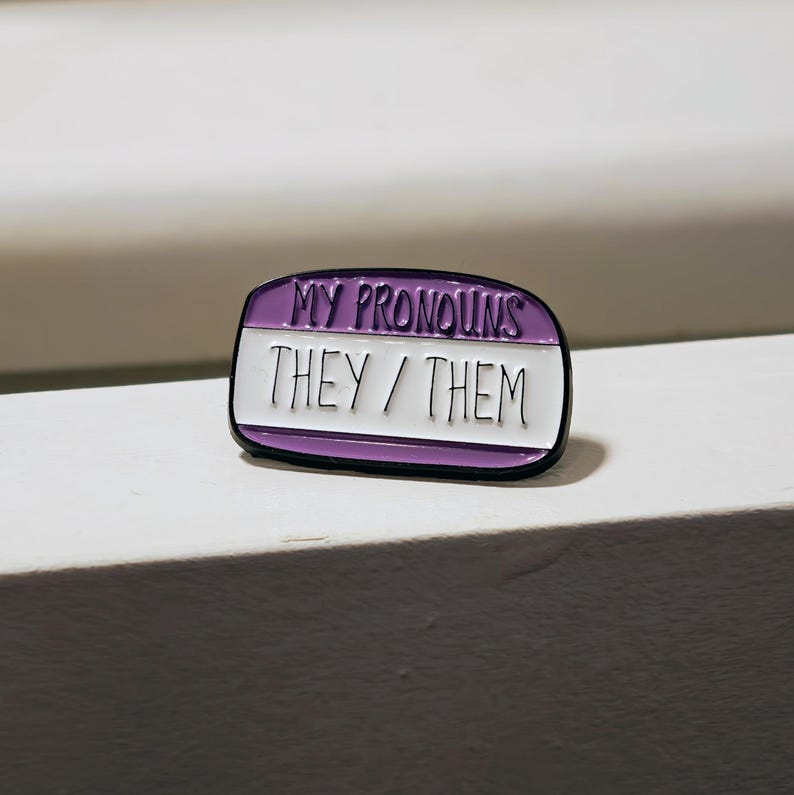 Puede incluir: Un pin de esmalte de forma ovalada con un borde negro. La secci&oacute;n superior es morada con el texto MY PRONOUNS en negro. La secci&oacute;n inferior es blanca con el texto THEY / THEM en negro, imitando la apariencia de una etiqueta de nombre.