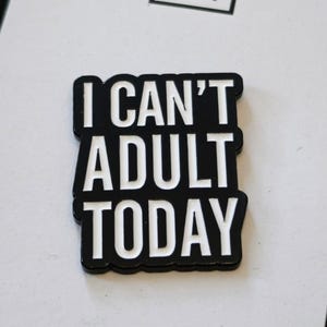 Puede incluir: Un pin de esmalte rectangular con fondo negro y letras blancas en negrita que dicen I CAN'T ADULT TODAY. El texto est&aacute; dispuesto en tres l&iacute;neas apiladas con un estilo de fuente sans-serif y un acabado suave y brillante en toda la pieza.