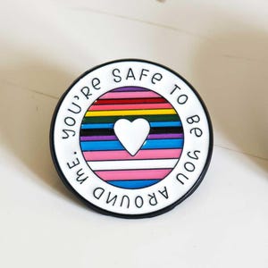 Puede incluir: Un pin redondo de esmalte con un borde blanco y el texto negro "YOU'RE SAFE AROUND ME". El centro contiene rayas horizontales en rojo, naranja, amarillo, verde, negro, marr&oacute;n, azul claro, blanco y rosa, con un coraz&oacute;n blanco en el centro.