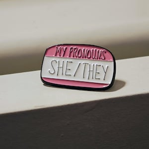 Puede incluir: Un pin de esmalte rectangular con esquinas redondeadas, que presenta una secci&oacute;n superior rosa con el texto negro MY PRONOUNS y una secci&oacute;n inferior blanca con el texto negro SHE/THEY. El pin tiene un borde negro fino que perfila la forma.