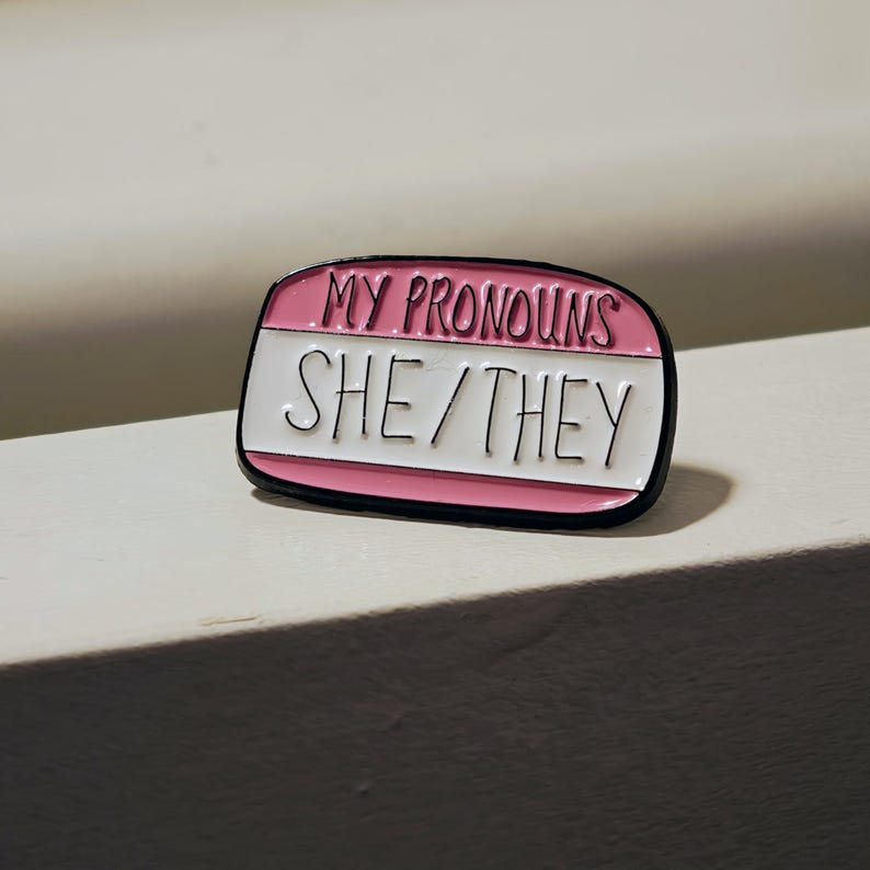 Puede incluir: Un pin de esmalte rectangular con esquinas redondeadas, que presenta una secci&oacute;n superior rosa con el texto negro MY PRONOUNS y una secci&oacute;n inferior blanca con el texto negro SHE/THEY. El pin tiene un borde negro fino que perfila la forma.