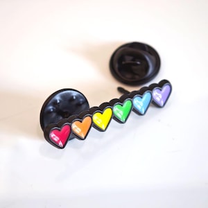 Puede incluir: Un pin de solapa de metal negro con una fila de seis corazones peque&ntilde;os en un degradado de arco&iacute;ris de esmalte rojo, naranja, amarillo, verde, azul y morado. El pin tiene dos cierres circulares negros situados detr&aacute;s de la barra de metal.