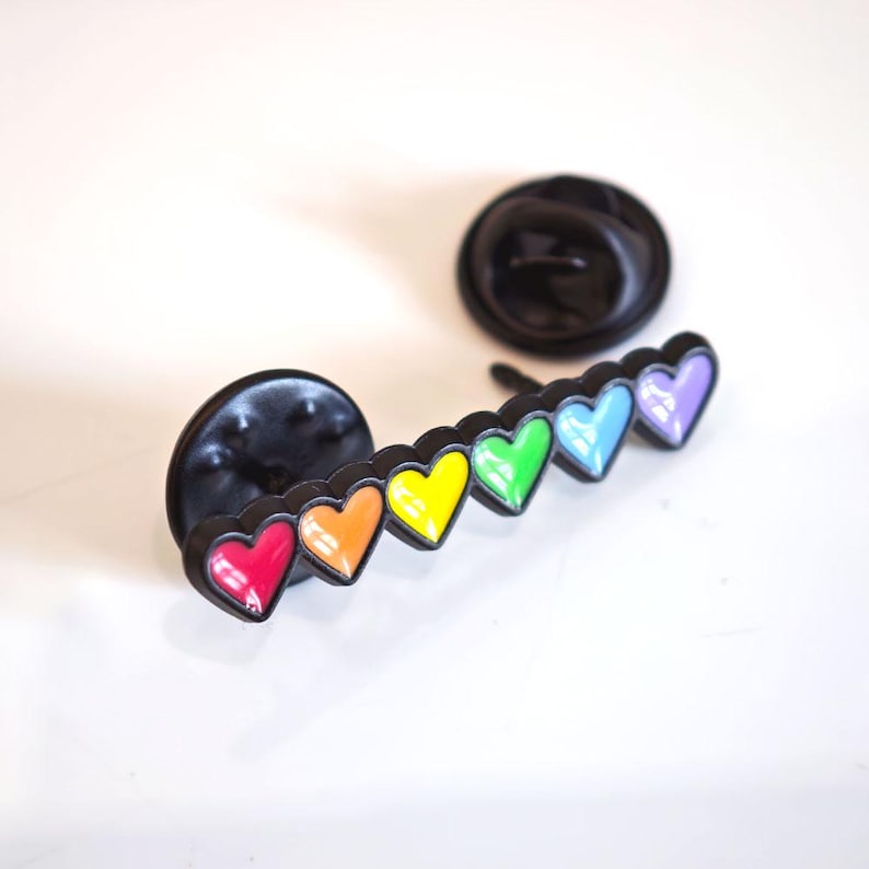 Puede incluir: Un pin de solapa de metal negro con una fila de seis corazones peque&ntilde;os en un degradado de arco&iacute;ris de esmalte rojo, naranja, amarillo, verde, azul y morado. El pin tiene dos cierres circulares negros situados detr&aacute;s de la barra de metal.