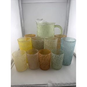 May include: A glass pitcher and ten matching tumblers featuring a textured, crackled finish in various translucent colors including yellow, orange, green, and blue. Each piece has a distinct web-like pattern across the exterior surface.