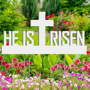 May include: A white metal garden stake with a cross design and the words "HE IS RISEN" in a garden bed filled with colorful flowers. The flowers include red, pink, purple, and yellow blooms, with green foliage in the background.