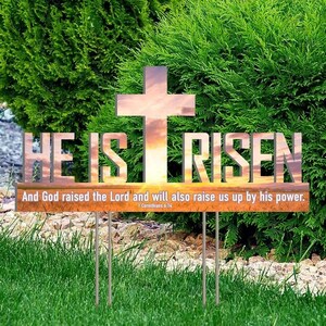 May include: A metal garden stake with the words "HE IS RISEN" and a cross design. The stake also includes the text "And God raised the Lord and will also raise us up by his power." The stake is placed in a grassy area with green bushes in the background.