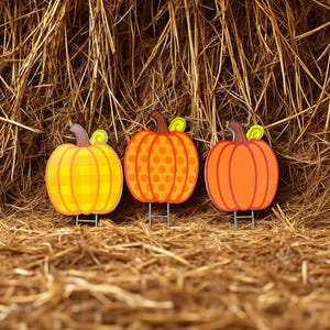 May include: Three decorative pumpkin yard stakes in yellow, orange, and red-orange. Each pumpkin has a brown stem and green curly vine. The pumpkins are set against a backdrop of hay.