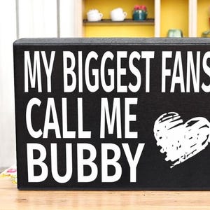 May include: Black wooden sign with white text that reads "My biggest fans call me Bubby" with a white heart graphic.