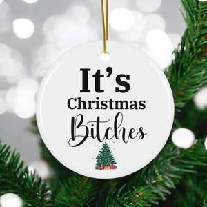 May include: A white ceramic Christmas ornament with the text "It's Christmas Bitches" in black. A small Christmas tree graphic is at the bottom. The ornament hangs from a gold string and is displayed on a Christmas tree.