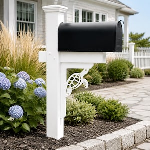 May include: A black metal mailbox mounted on a white post. The post features a decorative white bracket with a cutout design of a sea turtle. The mailbox is positioned in a garden setting with blue hydrangea flowers and green shrubs nearby.
