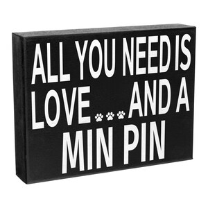 May include: Black wooden sign with white text that reads "All you need is love... and a Min Pin".