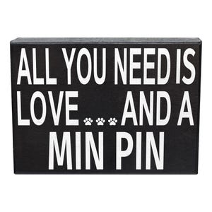 May include: Black wooden sign with white text that reads "All you need is love... and a Min Pin".