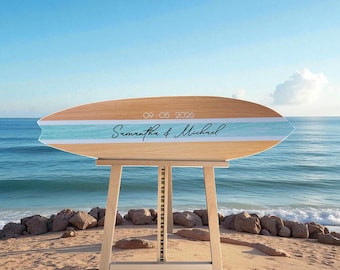 May include: A wooden surfboard-shaped sign resting on a light-colored wooden easel. The sign features a horizontal light blue stripe, the text 'Samantha & Michael' in black script, and the date '09.05.2026' printed in white.
