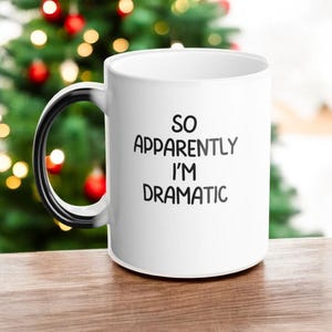 Könnte beinhalten: Weißer Keramikbecher mit schwarzem Henkel und dem Text "SO APPARENTLY I'M DRAMATIC" in Schwarz. Der Becher steht auf einer Holzoberfläche mit verschwommenem Hintergrund eines Weihnachtsbaums.