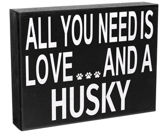 May include: A rectangular black wooden sign with white text that reads ALL YOU NEED IS LOVE... AND A HUSKY, featuring three small white paw prints between the words LOVE and AND. The sign has a clean, block-letter design on a solid black background.
