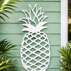 May include: A white, flat, cutout wall decoration shaped like a pineapple. The design features a detailed, open-work lattice pattern across the body of the fruit and pointed, stylized leaves at the top, creating a clean, graphic silhouette.