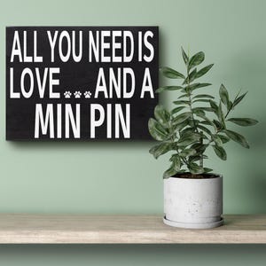 May include: Black wooden sign with white text that reads "All you need is love... and a Min Pin". The sign is hanging on a light green wall above a shelf with a potted plant.