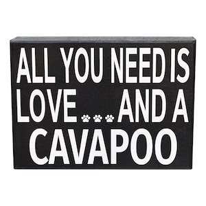 May include: A rectangular black wooden sign with white text that reads ALL YOU NEED IS LOVE... AND A CAVAPOO, featuring three small white paw prints between the words LOVE and AND, designed with a clean, bold, and high-contrast aesthetic.