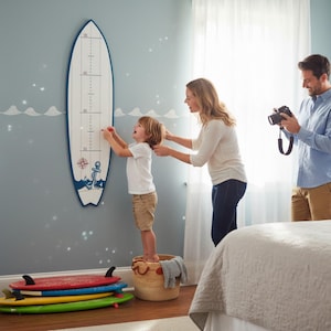 May include: A white and blue surfboard-shaped growth chart with nautical-themed illustrations. A child stands on a basket, marking their height. A parent assists, while another takes a photo. Several surfboards are stacked on the floor.