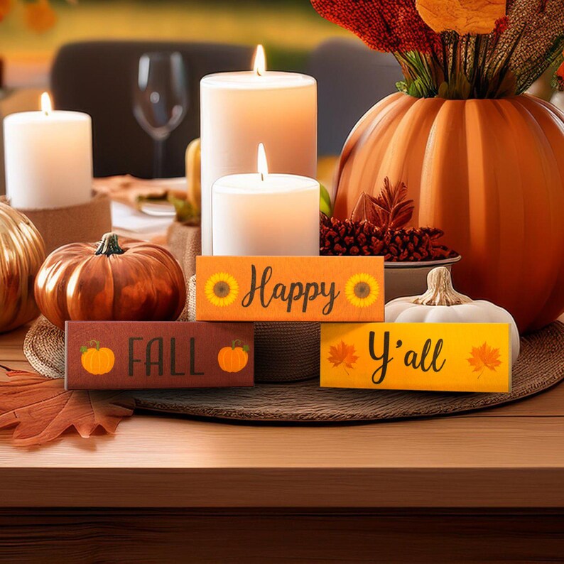 May include: Three wooden blocks stacked on top of each other. The top block is orange and says 'Happy' with a sunflower on each side. The middle block is maroon and says 'Fall' with a small orange pumpkin on each side. The bottom block is yellow and says 'Y'all' with a red maple leaf on each side. The blocks are sitting on a wooden shelf with a pumpkin and a gourd on either side of the blocks.