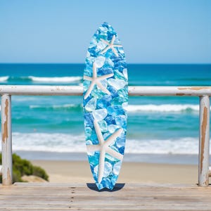 May include: A surfboard with a blue and white design featuring a pattern of blue glass and white starfish. The surfboard is leaning against a white railing with a beach and ocean in the background.
