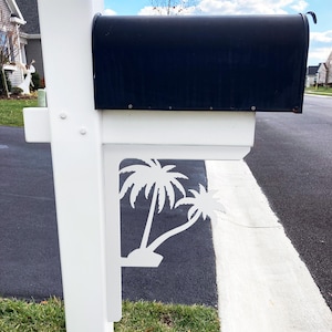 May include: A black mailbox with a white base and a decorative white palm tree design. The mailbox is mounted on a white post and is located near a paved road and green grass.