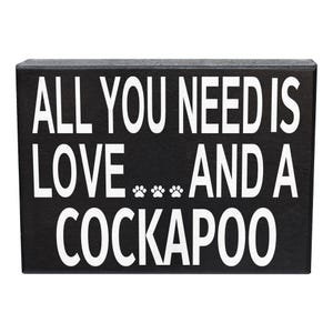 May include: A rectangular black wooden sign with white text that reads ALL YOU NEED IS LOVE... AND A COCKAPOO. The text includes three small white paw prints between the words LOVE and AND, featuring a clean, bold, sans-serif font style.