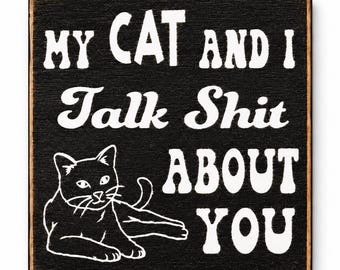 May include: A square black wooden sign with distressed edges featuring white text that reads MY CAT AND I Talk Shit ABOUT YOU and a white line art illustration of a cat lying down in the lower left corner of the sign surface.