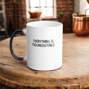 Puede incluir: Taza de café de cerámica blanca con asa negra. La taza tiene el texto "EVERYTHING IS FIGUREOUTABLE" impreso en negro en la parte delantera.