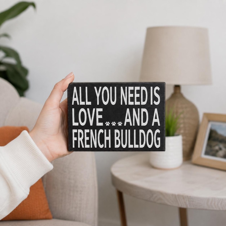 A hand holds a rectangular black sign with white text that reads 'ALL YOU NEED IS LOVE.