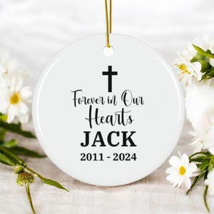 Può includere: Ornamento commemorativo rotondo in ceramica bianca con cordino dorato per appenderlo. Presenta una croce nera e la scritta "Forever in Our Hearts JACK 2011-2024" in corsivo. Fiori bianchi sullo sfondo.
