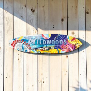 May include: A colorful surfboard-shaped wooden sign with the word "Wildwoods" in white lettering. The sign features a vibrant illustration of a Ferris wheel, beach balls, and a bus. The sign is mounted on a weathered, light-colored wooden wall.